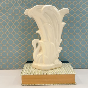 May include: A glossy off-white ceramic vase featuring a sculptural design with layered, petal-like vertical ridges and a swan-shaped base. The vase is positioned on top of a rectangular book with a patterned cover and a thick, tan-colored page edge.