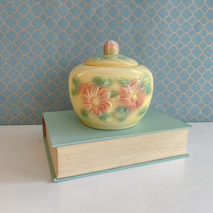 Puede incluir: Un azucarero vintage de cerámica amarilla con tapa, adornado con diseños florales rosas y verdes. El azucarero está sobre un libro azul claro, sobre un fondo azul estampado.