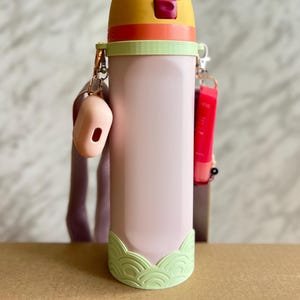 May include: A pastel pink water bottle with a yellow and orange lid. The bottle has a green wave-like design at the base and is accessorized with a pink case and a red lip gloss. The bottle is approximately 23 cm tall.