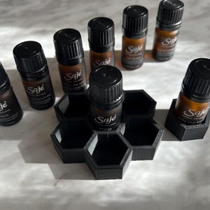 May include: A collection of small brown glass essential oil bottles with black caps, labeled "Saje", arranged around a black hexagonal holder. The bottles are filled with a clear liquid. The background is a light-colored surface.