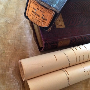 May include: A close-up of a vintage music player roll with the title "Bostonian March" on a brown book with the word "Eugene" on the cover. The roll is on top of a beige fabric.