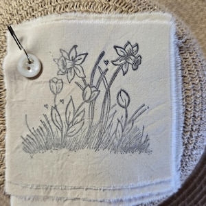 May include: A hand-stitched fabric patch with a black and white line drawing of daffodils and buds on a beige background.
