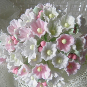May include: A close-up of a bouquet of delicate, handmade flowers. The flowers are primarily white and pink, with small yellow centers. The arrangement includes small pink and green accents, creating a soft, pastel color palette. The flowers appear to be made of paper or fabric.