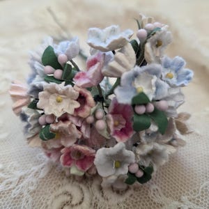 May include: A small bouquet of paper flowers in shades of pink, white, and blue. The flowers have green leaves and small pink berry accents. The bouquet is set against a lace background.