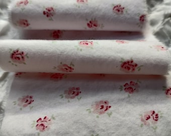 May include: Close-up of white fabric with a repeating pattern of small pink roses. The fabric is folded into multiple layers, creating a soft and delicate look.