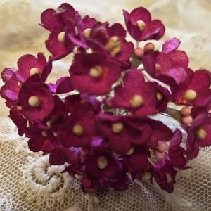 May include: A cluster of deep magenta paper flowers with yellow centers and small pink buds. The flowers are arranged on a light-colored, textured background, creating a delicate and vintage aesthetic.