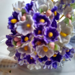 May include: A cluster of small, delicate paper flowers in shades of purple, white, and yellow. The flowers are arranged in a bouquet and have a vintage aesthetic.