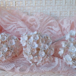 May include: Three clusters of small white artificial flowers with a delicate, slightly ruffled texture. The flowers are arranged on a pink lace background.