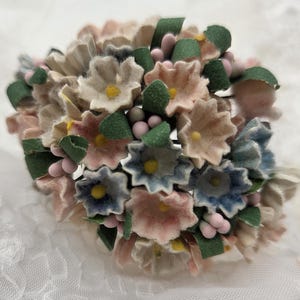 May include: A cluster of handmade paper flowers in shades of pink, blue, and beige, with yellow centers and green leaves. The arrangement includes small pink and gray bead-like accents, creating a delicate floral design.
