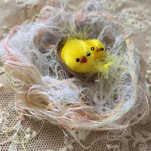 May include: A handcrafted nest made of woven brown material, holding two fluffy yellow chicks with black eyes and red beaks. The nest is surrounded by a delicate, multi-colored yarn, set against a textured, cream-colored background.