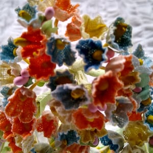 May include: A cluster of crocheted flowers in various colors, including blue, pink, white, and orange. The flowers are arranged in a circular pattern and have a delicate, lace-like texture.