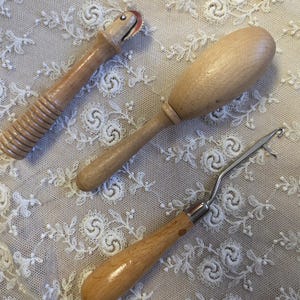 May include: Three wooden tools for crafting, including a roller with a red wheel, a rounded handle tool, and a hook tool with a wooden handle and metal hook. The tools are displayed on a white lace fabric.