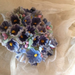 May include: A bouquet of blue and purple fabric flowers with yellow centers and pink accents. The flowers are arranged in a circular shape and are surrounded by a white fabric.