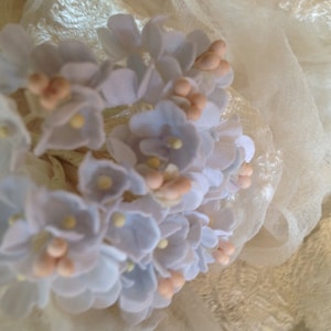 May include: A cluster of small, pale blue fabric flowers with pink centers. The flowers are arranged in a bouquet and are sitting on a white fabric background.