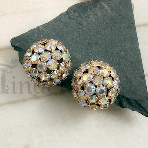 May include: A pair of spherical earrings featuring a dense cluster of round, iridescent, light-reflecting crystals set in a gold-toned metal frame. Each earring is shaped like a small, shimmering dome covered in faceted stones that display rainbow hues.
