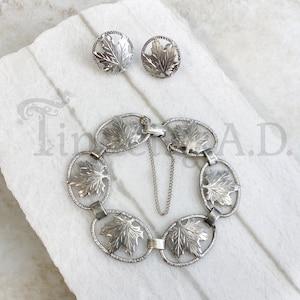 May include: A silver bracelet with five oval-shaped links, each featuring a detailed maple leaf design. The bracelet has a silver clasp and chain. Two matching silver earrings with maple leaf designs are also included.