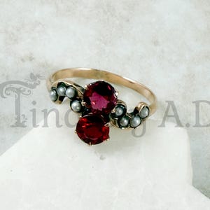 May include: A gold ring featuring two round, deep red gemstones and small, gray pearls. The ring's band is a warm gold color, and the gemstones are set in a cluster design. The pearls are arranged around the gemstones.