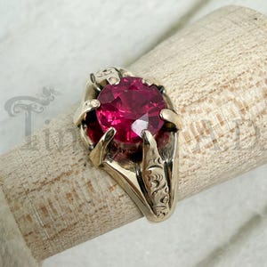 May include: A gold ring featuring a large, round, faceted deep pink gemstone set in a prong setting. The metal band displays intricate, engraved scrollwork patterns along the sides, providing a detailed texture to the gold surface.