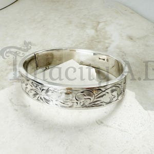 May include: A silver-colored hinged bangle bracelet with intricate floral and scrollwork engravings. The bracelet has a polished finish and a secure clasp. The word "Sterling" is stamped on the inside of the bracelet.