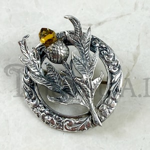 May include: A silver-toned brooch featuring a detailed thistle design. The brooch has a circular frame with floral detailing, surrounding a thistle flower and leaves. A faceted, yellow gemstone accents the top of the thistle.