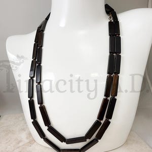 May include: A two-strand necklace featuring dark brown rectangular beads. The beads are strung together, creating a layered effect. The necklace is displayed on a white mannequin bust, showcasing its design and length. The beads have a polished finish.