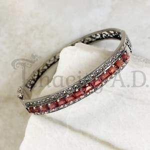 May include: A silver bangle bracelet with a delicate filigree design and a row of red square-shaped gemstones.