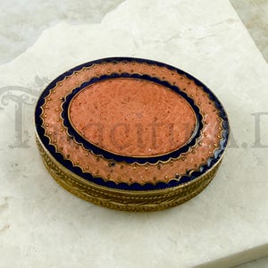 May include: An antique, oval-shaped trinket box with a coral-colored center and a decorative border. The box features a gold-colored metal base and a blue and peach enamel design. The lid has a scalloped edge.