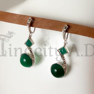 May include: A pair of silver-toned earrings with green gemstones. Each earring features a round, polished green stone at the bottom, a square green stone in the middle, and a decorative top. The earrings are displayed against a neutral background.