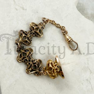 May include: A gold-toned antique watch chain with an ornate, floral design. The chain features a series of decorative links, a small chain, and a clasp. The seal at the end has a circular design.