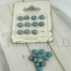 May include: A collection of small, round, light blue buttons with gold-toned metal edges. Eleven buttons are attached to a vintage off-white cardboard display card labeled La Mode, Superior Quality, and Czechoslovakia. Nine loose buttons are below.