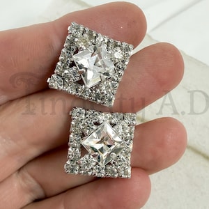 May include: A pair of square-shaped silver-tone earrings featuring a central princess-cut clear rhinestone surrounded by a border of smaller round clear rhinestones. The earrings have a metallic setting and a reflective, faceted surface design.
