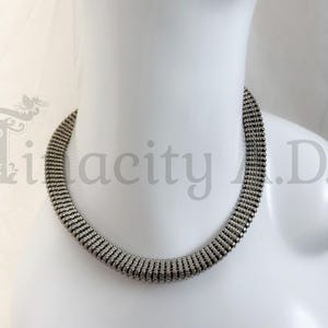 May include: A gunmetal-toned necklace featuring a wide, flexible mesh band composed of multiple rows of small, textured metallic links. The necklace has a uniform, cylindrical shape that rests against the neck, displaying a dark, reflective finish.