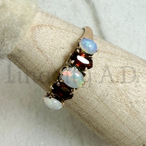 May include: A gold ring featuring an alternating row of three oval-shaped opals and two dark red oval-shaped garnets. The stones are set in a prong setting along the band, showcasing the iridescent white of the opals and the deep red of the garnets.