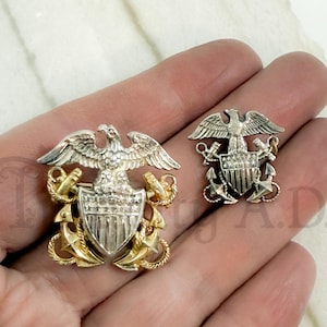 以下が含まれることがあります： 2つのヴィンテージミリタリー記章ピン。大きなピンは、金色のアンカーとアクセントが付いたシールドの上に銀色のワシが描かれています。小さなピンも同様のデザインです。どちらのピンも金属製である可能性があります。