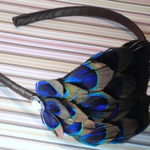 May include: A brown headband with a peacock feather embellishment. The feathers are blue, green, and brown. A silver-toned button is attached to the headband.