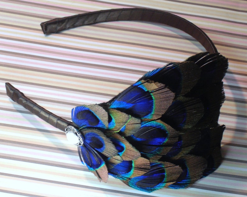 May include: A brown headband with a peacock feather embellishment. The feathers are blue, green, and brown. A silver-toned button is attached to the headband.
