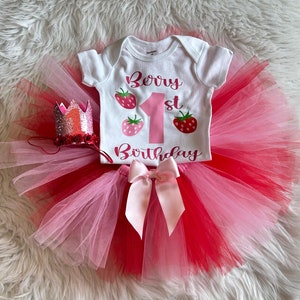Puede incluir: Un tut&uacute; de tul rosa y rojo con un body blanco que dice "Berry 1st Birthday" y un lazo rosa. El body tiene tres gr&aacute;ficos de fresas. Una corona de purpurina rosa y plateada est&aacute; en el lado izquierdo de la imagen.