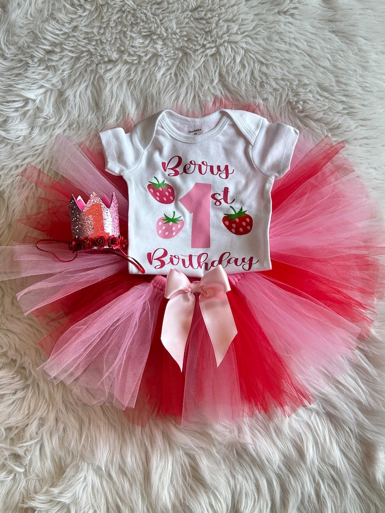 Puede incluir: Un tut&uacute; de tul rosa y rojo con un body blanco que dice "Berry 1st Birthday" y un lazo rosa. El body tiene tres gr&aacute;ficos de fresas. Una corona de purpurina rosa y plateada est&aacute; en el lado izquierdo de la imagen.