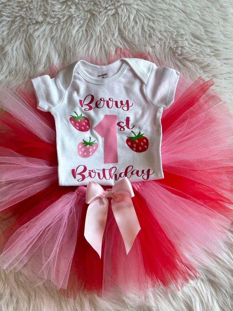 Puede incluir: Un body blanco con el texto "Berry 1st Birthday" en letras rosas y gr&aacute;ficos de fresas. El body se combina con un tut&uacute; de tul rosa y rojo con un lazo de sat&eacute;n rosa.