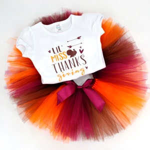 May include: A white onesie with the text "Lil Miss Thanks Giving" and a small turkey graphic. The onesie is paired with a multi-colored tulle skirt with brown, orange, and burgundy layers.