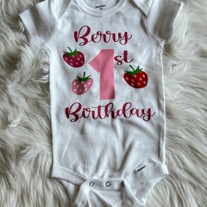 Puede incluir: Un body blanco para beb&eacute; con el texto "Berry 1st Birthday" en letras rosas. El n&uacute;mero 1 es grande y rosa. Hay tres gr&aacute;ficos de fresas rojas y rosas alrededor del n&uacute;mero 1.
