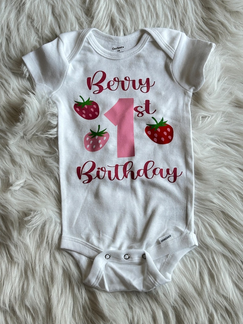Puede incluir: Un body blanco para beb&eacute; con el texto "Berry 1st Birthday" en letras rosas. El n&uacute;mero 1 es grande y rosa. Hay tres gr&aacute;ficos de fresas rojas y rosas alrededor del n&uacute;mero 1.