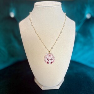 Puede incluir: Un collar dorado con un colgante de concha. El colgante es una concha rosa y blanca. El collar tiene pequeñas perlas blancas espaciadas a lo largo de la cadena. El collar se exhibe en un maniquí blanco sobre un fondo verde azulado.