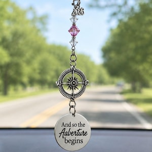 May include: A silver car charm with a compass design, pink crystals, and the year 2023. The charm also has a circular pendant with the phrase "And so the Aabenture begins". The charm hangs from a car's rearview mirror.