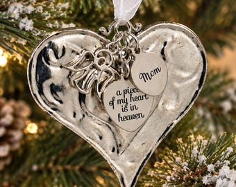 May include: A silver-toned heart-shaped ornament hanging from a white ribbon. It features a small angel charm and two heart-shaped tags. One tag reads "Mom" and the other is engraved with the text "a piece of my heart is in heaven."