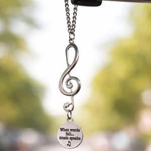 May include: Silver-colored treble clef car charm hanging from a chain. A small, round pendant at the bottom reads "When words fail... music speaks" with a musical note symbol. The background is blurred, suggesting an outdoor setting.