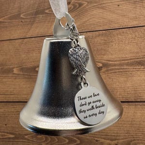 May include: A silver-toned metal bell ornament featuring a white ribbon loop at the top. Attached to the bell is a silver charm shaped like angel wings and a round silver disc engraved with the text: Those we love don't go away, they walk beside us every day.