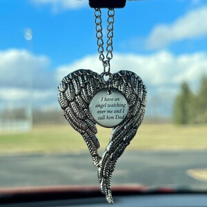 May include: Silver-colored angel wing car charm. The charm is shaped like a heart with detailed feathering. A circular pendant in the center reads, "I have an angel watching over me and I call him Dad."