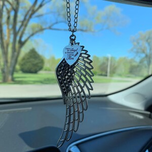 May include: Silver car charm with a heart-shaped pendant that reads "I have an Angel in Heaven I call her Mom". The charm is attached to a silver chain and has a silver angel wing design.