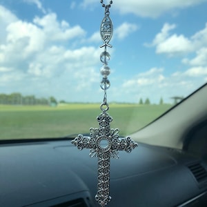 May include: A silver car charm featuring a cross with intricate details and a fish symbol with the word "JESUS" inscribed. The charm is hanging from a rearview mirror with white beads.